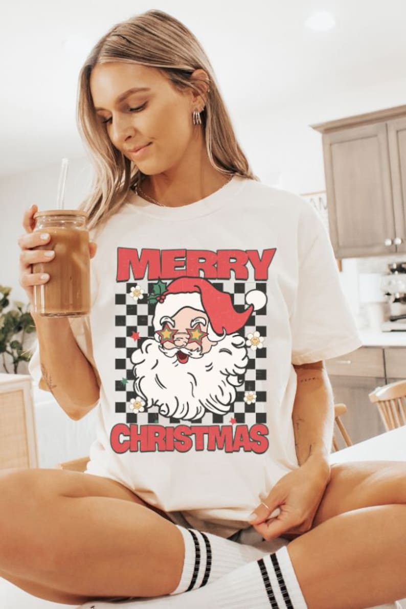 K&ouml;nnte beinhalten: Wei&szlig;es T-Shirt mit einem rot-wei&szlig; karierten Muster und einem Cartoon-Weihnachtsmann mit Sonnenbrille. Der Text "Merry Christmas" ist auf dem Shirt aufgedruckt.