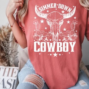 May include: A red t-shirt with a white graphic of a cow skull with horns, cactus plants, and stars. The text "SIMMER DOWN COWBOY" is printed on the shirt.