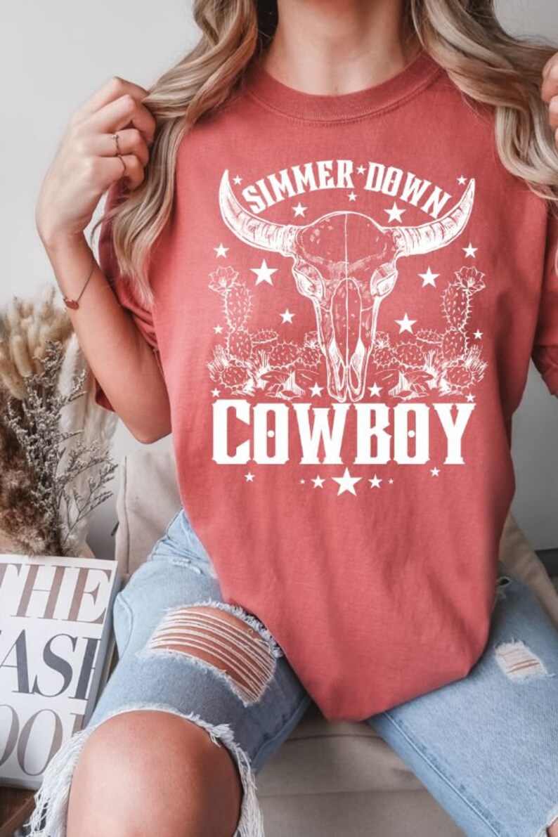 May include: A red t-shirt with a white graphic of a cow skull with horns, cactus plants, and stars. The text "SIMMER DOWN COWBOY" is printed on the shirt.