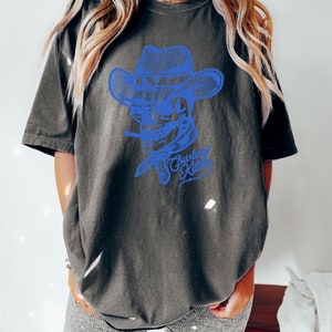 May include: A dark grey t-shirt featuring a blue graphic of a skull wearing a cowboy hat and a bandana. The text "Cowboy Killer" is beneath the skull.