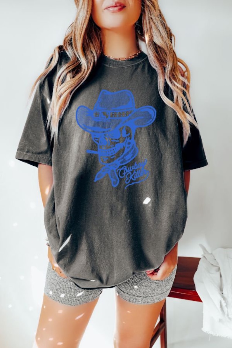 May include: A dark grey t-shirt featuring a blue graphic of a skull wearing a cowboy hat and a bandana. The text "Cowboy Killer" is beneath the skull.