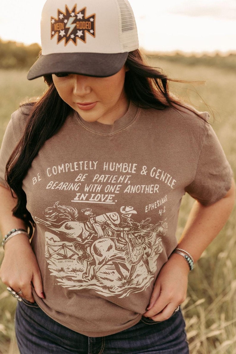 K&ouml;nnte beinhalten: Ein braunes T-Shirt mit einer Vintage-Illustration von Cowboys, die auf Pferden reiten. Das Shirt tr&auml;gt den Text "BE COMPLETELY HUMBLE & GENTLE BE PATIENT, BEARING WITH ONE ANOTHER IN LOVE. EPHESIANS 4:2".