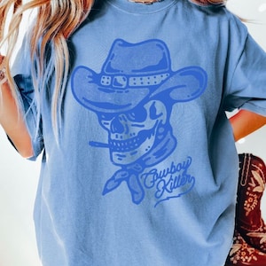 May include: A light blue t-shirt featuring a blue graphic of a skull wearing a cowboy hat and bandana. The text "Cowboy Killer" is written beneath the skull.