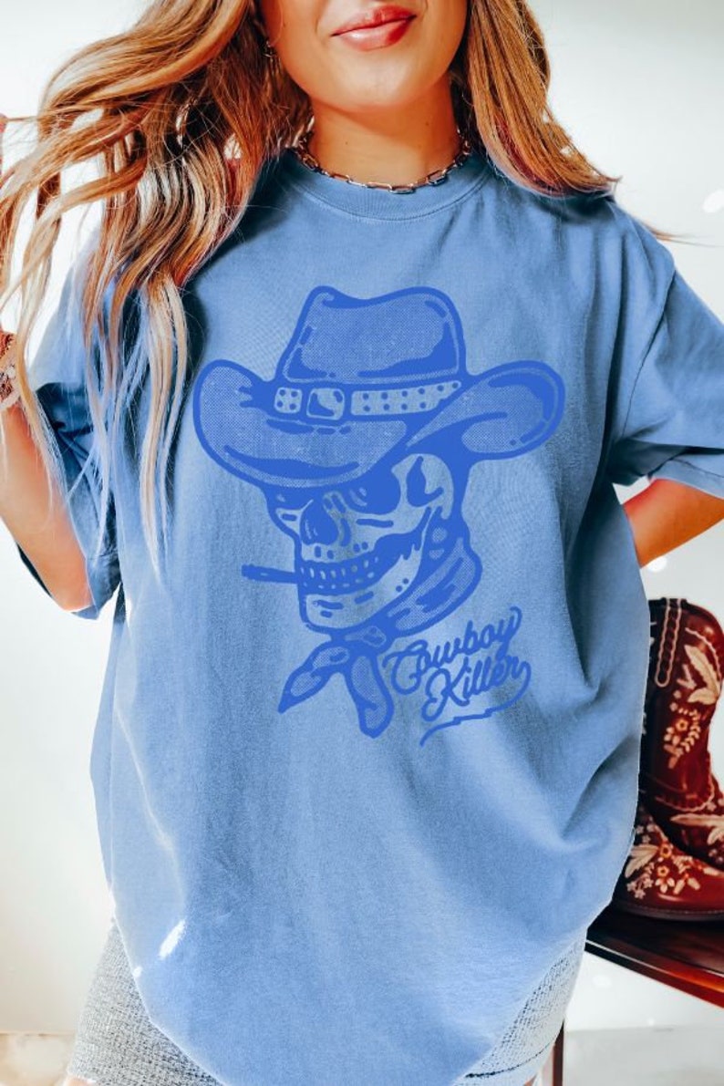 May include: A light blue t-shirt featuring a blue graphic of a skull wearing a cowboy hat and bandana. The text "Cowboy Killer" is written beneath the skull.