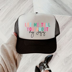 May include: A white trucker hat with a black mesh back. The front of the hat has the text "Let me tell you about my Jesus" in colorful letters.