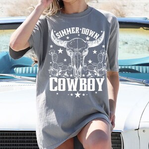 May include: A gray oversized t-shirt with a white graphic of a bull skull with horns, cactus, and stars. The text "SIMMER DOWN COWBOY" is printed on the shirt.