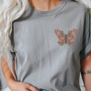 Puede incluir: Una camiseta gris con un dise&ntilde;o de mariposa floral en la parte delantera. La mariposa est&aacute; hecha de flores rosas y verdes.