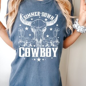 May include: A blue t-shirt with a white graphic of a cow skull with horns, cacti, and stars. The text "Summer Down Cowboy" is printed below the skull.