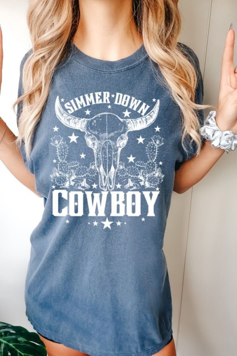 May include: A blue t-shirt with a white graphic of a cow skull with horns, cacti, and stars. The text "Summer Down Cowboy" is printed below the skull.