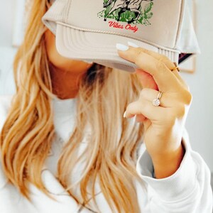 May include: A beige trucker hat with a cowgirl riding a horse graphic and the text "Cowgirl Vibes Only".