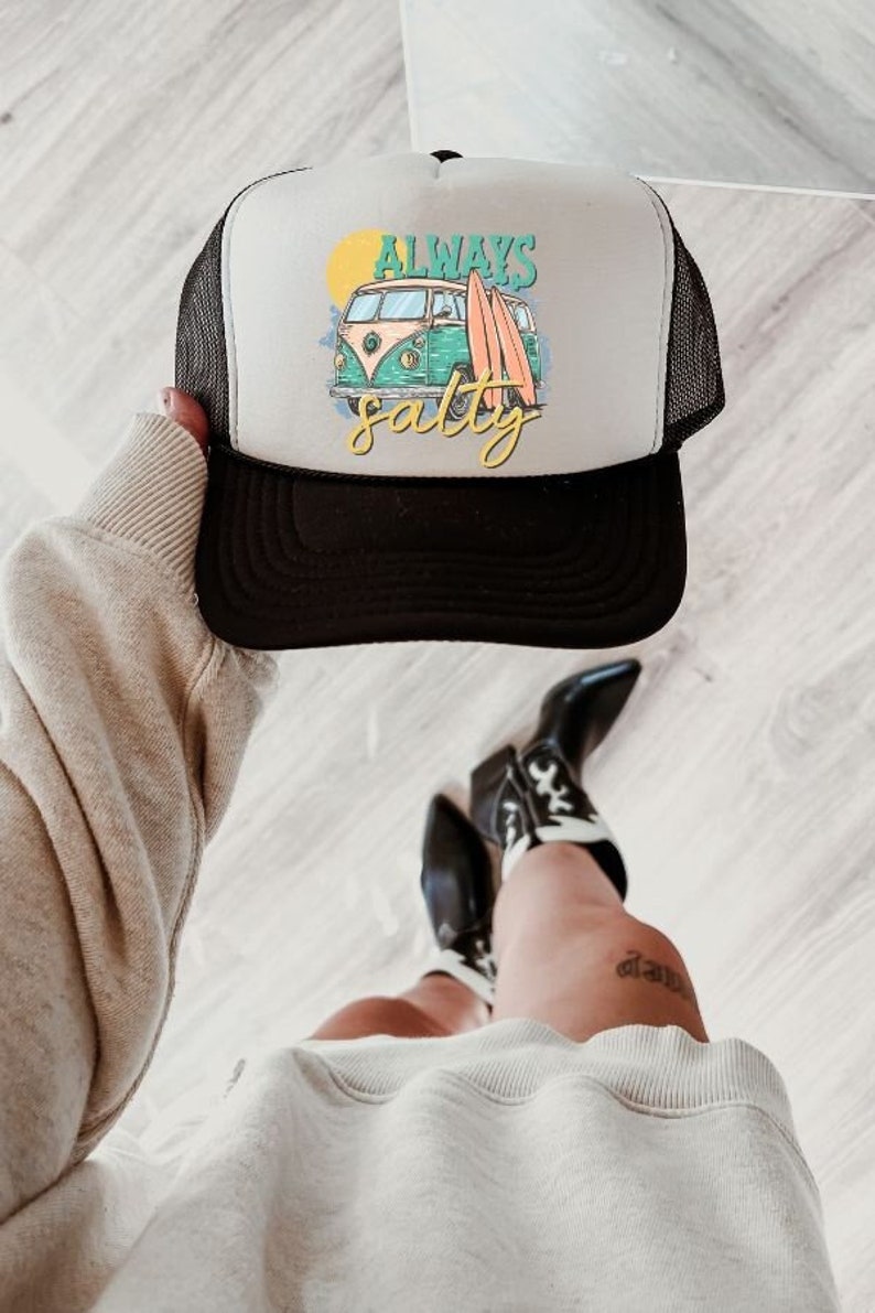 May include: A white and black trucker hat with a black mesh back. The front of the hat features a colorful illustration of a vintage van with surfboards and the text "Always Salty".