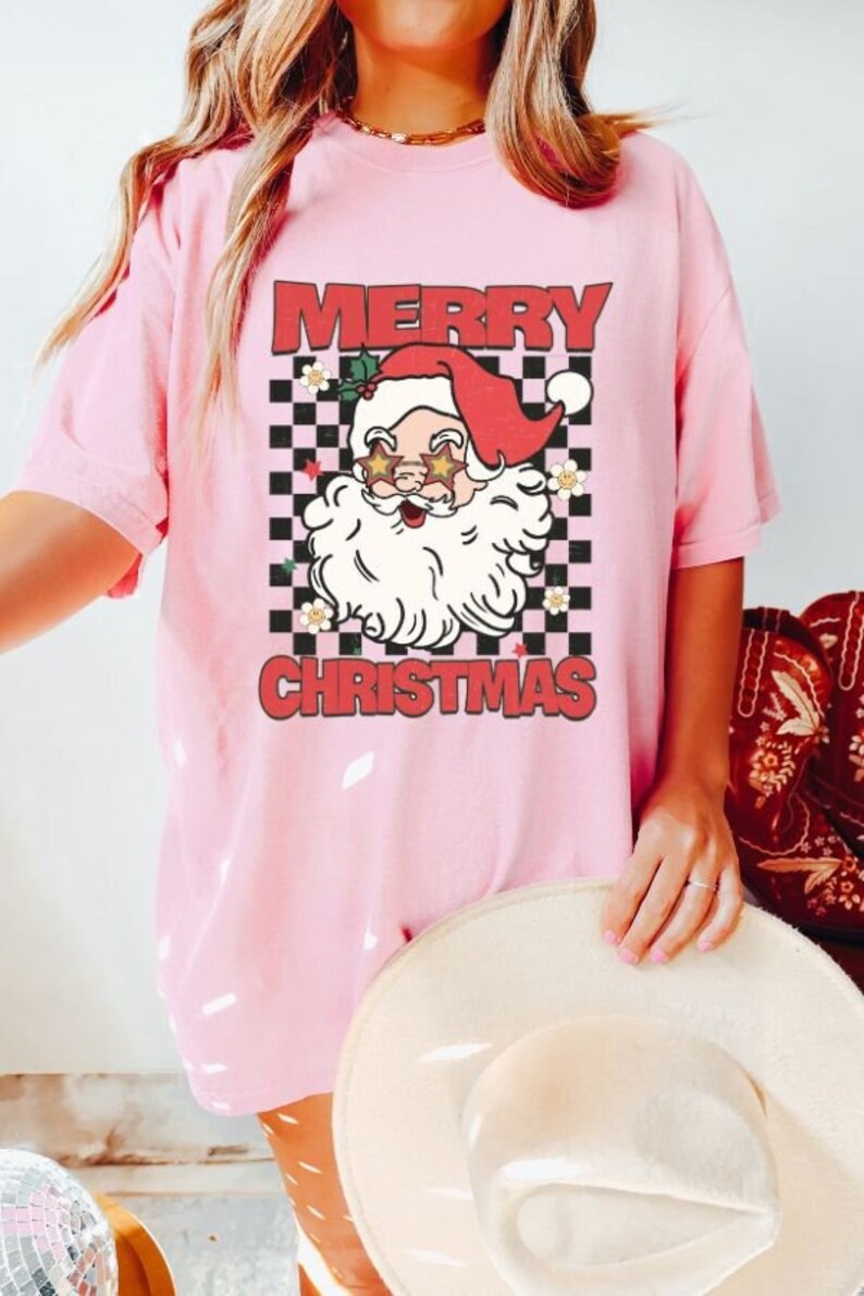 K&ouml;nnte beinhalten: Rosa T-Shirt mit kariertem Hintergrund und einem Comic-Weihnachtsmann mit Sonnenbrille. Der Text "Merry Christmas" ist auf dem Shirt aufgedruckt.
