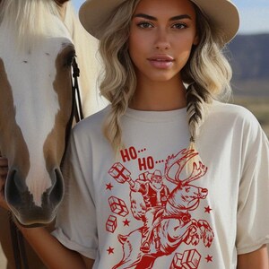 May include: A woman wearing a beige t-shirt with a red graphic of Santa Claus riding a reindeer and the text "HO...HO...HOWDY!" printed on it. She is wearing a beige hat and is standing next to a white horse.