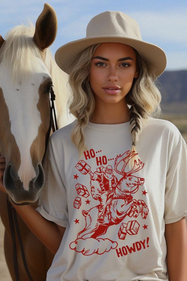 May include: A woman wearing a beige t-shirt with a red graphic of Santa Claus riding a reindeer and the text "HO...HO...HOWDY!" printed on it. She is wearing a beige hat and is standing next to a white horse.