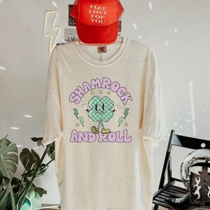 May include: A cream-colored t-shirt with a shamrock graphic and the text "Shamrock and Roll" in pink. A red mesh baseball cap with the text "Mad Love For You" is on top of the shirt.