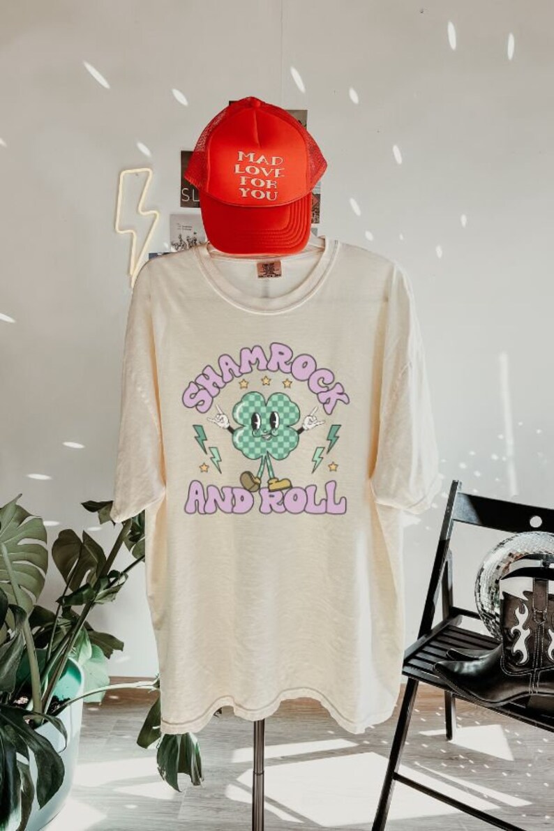 May include: A cream-colored t-shirt with a shamrock graphic and the text "Shamrock and Roll" in pink. A red mesh baseball cap with the text "Mad Love For You" is on top of the shirt.