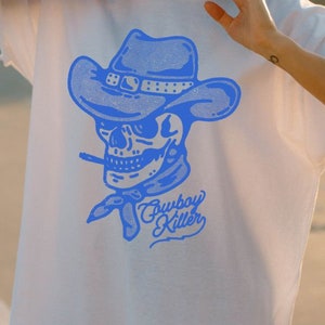 May include: A white t-shirt featuring a blue graphic of a skull wearing a cowboy hat and the text "Cowboy Killer".