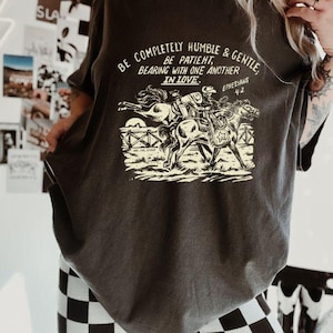 K&ouml;nnte beinhalten: Ein dunkelgraues T-Shirt mit einem grafischen Aufdruck von zwei Personen, die auf Pferden reiten. Der Text auf dem Shirt lautet "Be completely humble & gentle. Be patient, bearing with one another in love. Ephesians 4:2"