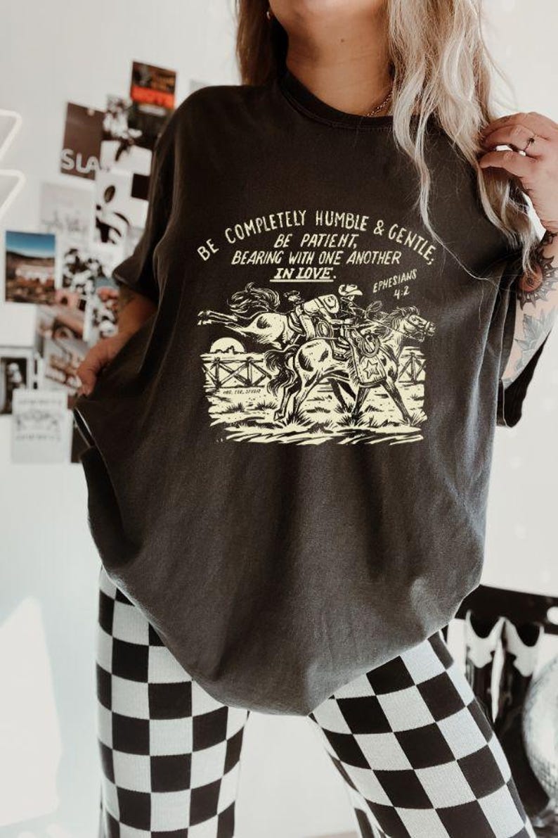K&ouml;nnte beinhalten: Ein dunkelgraues T-Shirt mit einem grafischen Aufdruck von zwei Personen, die auf Pferden reiten. Der Text auf dem Shirt lautet "Be completely humble & gentle. Be patient, bearing with one another in love. Ephesians 4:2"