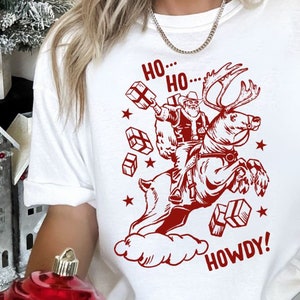 May include: White t-shirt with a red graphic of Santa Claus riding a reindeer and holding presents. The text "Ho... Ho... Howdy!" is printed on the shirt.