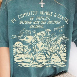 K&ouml;nnte beinhalten: Tealblaues T-Shirt mit einer Grafik im Vintage-Stil von zwei Pferden und einem Reiter. Der Text auf dem Shirt lautet "Be completely humble & gentle: Be patient, bearing with one another in love. Ephesians 4:2"
