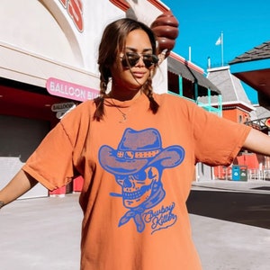 May include: A woman wearing a burnt orange t-shirt with a blue graphic of a skull wearing a cowboy hat and bandana. The text "Cowboy Killer" is printed below the skull.