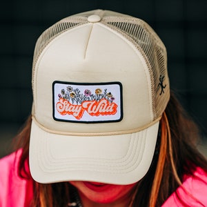 May include: A beige trucker hat with a black mesh back. The hat has a patch on the front that says "Stay Wild" in orange letters with a white outline. The patch also features a small illustration of flowers.