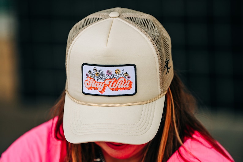 May include: A beige trucker hat with a black mesh back. The hat has a patch on the front that says "Stay Wild" in orange letters with a white outline. The patch also features a small illustration of flowers.