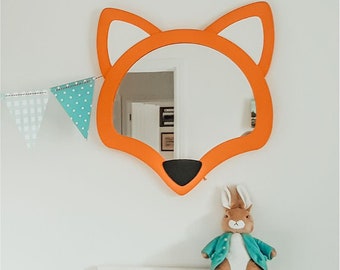 May include: Orange fox-shaped mirror with a black nose and a reflection of a room in the background. A small stuffed rabbit wearing a blue shirt is in the foreground.