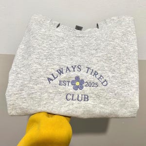 May include: A grey marl sweatshirt featuring navy blue embroidered text reading "ALWAYS TIRED CLUB EST 2025". A small flower graphic with a yellow centre is between "EST" and "2025".