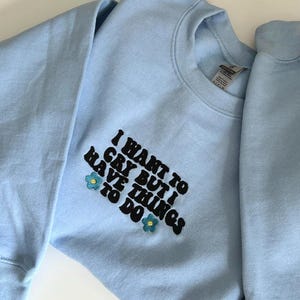 May include: A pale blue sweatshirt featuring the embroidered phrase "I WANT TO CRY BUT I HAVE THINGS TO DO" in black, with floral accents. The garment is made of a soft fabric.