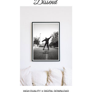 May include: Black and white photograph of a person skateboarding down a street in the city. The person is wearing a jacket and pants and is performing a trick on the skateboard. The photo is framed in a black frame.