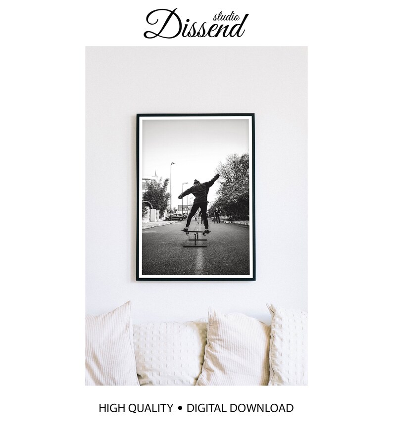 May include: Black and white photograph of a person skateboarding down a street in the city. The person is wearing a jacket and pants and is performing a trick on the skateboard. The photo is framed in a black frame.
