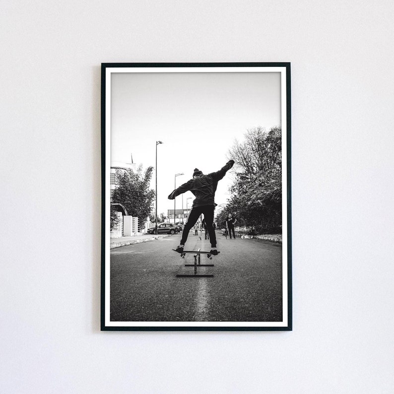 May include: Black and white photograph of a person skateboarding on a ramp in the street. The person is wearing a jacket and jeans. There are trees and buildings in the background.