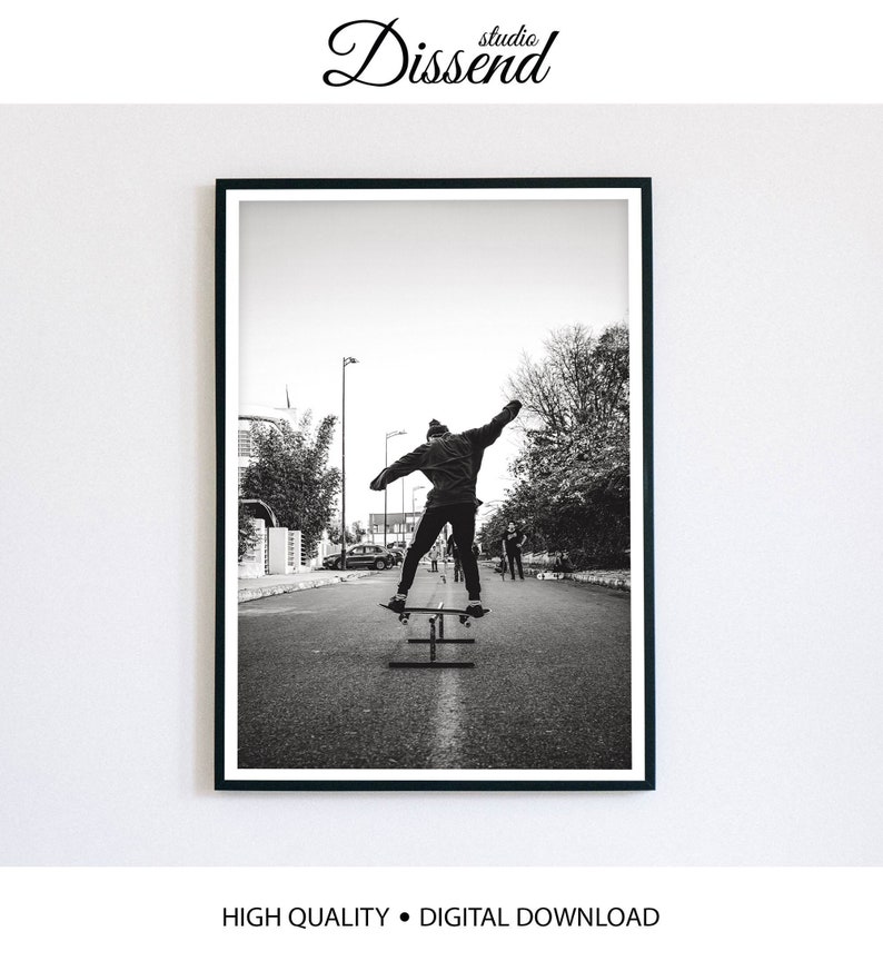 May include: Black and white photograph of a person skateboarding down a street. The person is wearing a jacket and jeans and is in mid-air. There are trees and buildings in the background.