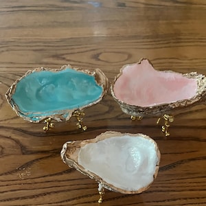 May include: Three decorative oyster shell dishes featuring smooth, glossy interiors in turquoise, light pink, and white. Each shell has a metallic gold-painted rim and is supported by three small, ornate gold-toned metal legs for elevation.