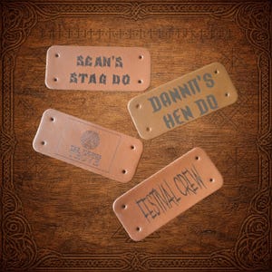 May include: Four rectangular leather tags with text. One tag reads "SEAN'S STAG DO.", another says "DANNIT'S HEN DO", a third says "THE VIKINGS TROVE", and the last says "FESTIVAL CREW". The tags are tan and brown, with dark grey text.