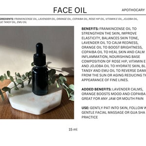 May include: A 15 ml bottle of face oil with a black dropper. The bottle is sitting on a white marble surface with green leaves. The label lists the ingredients and benefits of the oil, including frankincense, lavender, orange, copaiba, rose hip, vitamin E, jojoba, blue tansy, and emu oil.