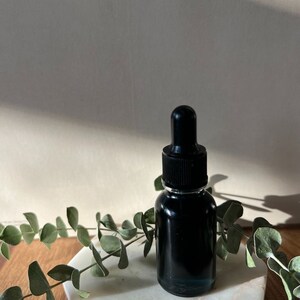 May include: A dark glass bottle with a black dropper lid, filled with a dark liquid, sits on a white marble hexagon-shaped coaster. The coaster is on a wooden surface with green leafy branches around the bottle.