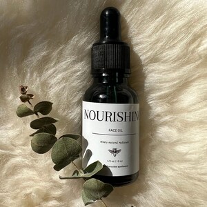 May include: A small, dark glass bottle with a black dropper top. The bottle is labelled "Nourishing Face Oil" and "renew natural radiance". The label also includes a small bee illustration and the text "1/2 oz | 15 ml" and "honeybee apothecary".