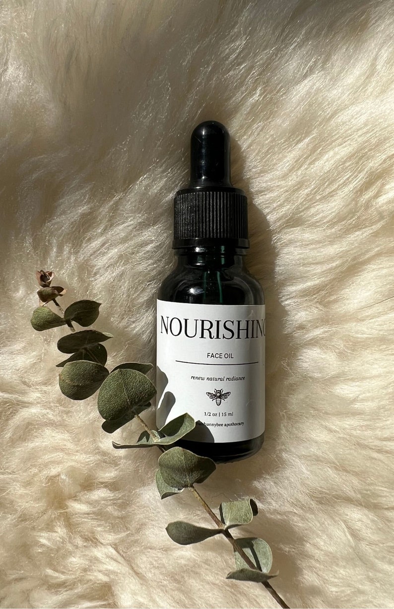 May include: A small, dark glass bottle with a black dropper top. The bottle is labelled "Nourishing Face Oil" and "renew natural radiance". The label also includes a small bee illustration and the text "1/2 oz | 15 ml" and "honeybee apothecary".