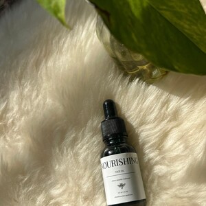 May include: A small, dark brown glass bottle with a dropper top. The bottle is labelled "Nourishing Face Oil" and contains 1.2 fl oz (35 ml) of oil. The bottle is sitting on a white fluffy surface.