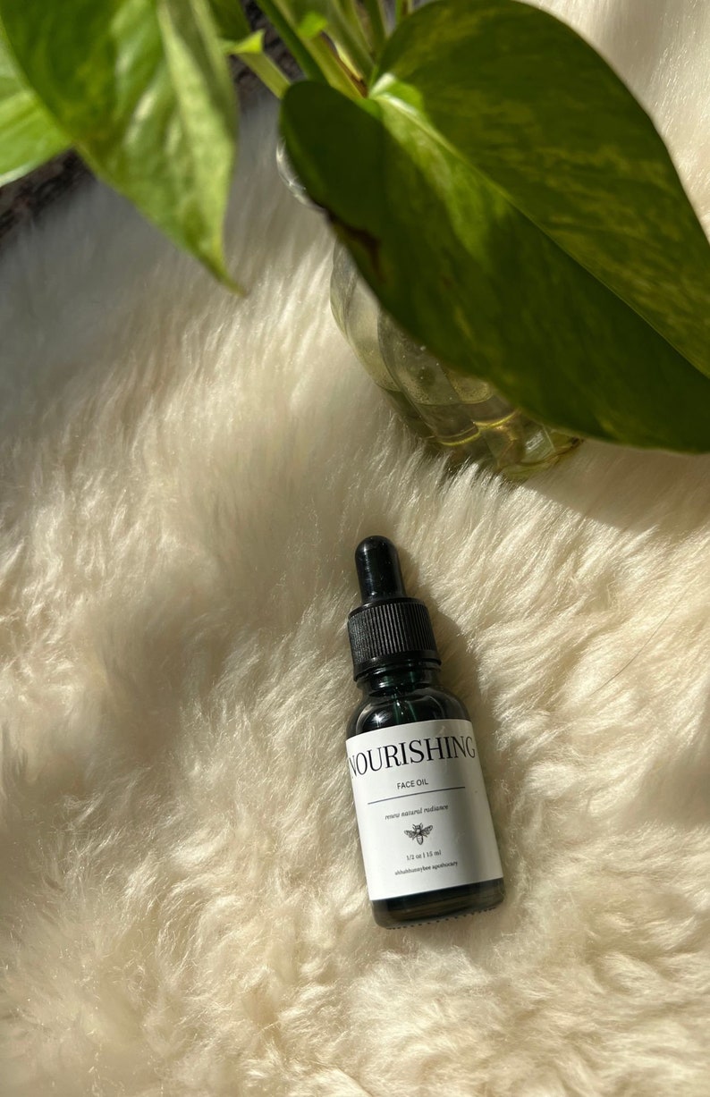 May include: A small, dark brown glass bottle with a dropper top. The bottle is labelled "Nourishing Face Oil" and contains 1.2 fl oz (35 ml) of oil. The bottle is sitting on a white fluffy surface.