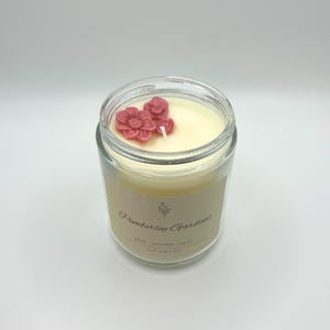 May include: A clear glass jar candle with a cream-colored wax and three pink flower-shaped wax decorations. The label reads "Pemberley Gardens" with "Rose, Jasmine, Lilac" and "Love and Loss" below. The candle is on a white background.