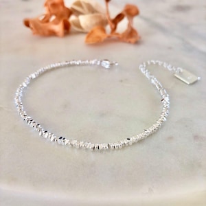 Peut inclure: Un bracelet argenté délicat avec un design perlé texturé. Le bracelet présente de petites perles à facettes qui captent la lumière, créant un effet scintillant. Le fermoir et une petite étiquette sont également visibles. Le bracelet est présenté sur une surface en marbre.