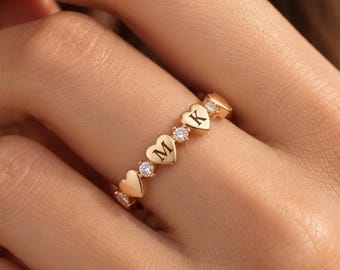 May include: A gold band ring featuring a series of alternating heart-shaped charms and small clear round stones. Two of the heart charms are engraved with the letters M and K, while the other hearts remain plain, creating a repeating pattern.