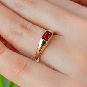 Op de afbeelding: Een gouden ring met een rode rechthoekige edelsteen. De ring zit aan een vinger.