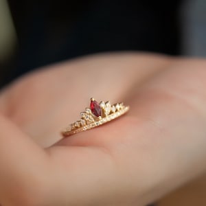 May include: A gold ring with a red gemstone in the center, surrounded by smaller clear gemstones. The ring is shaped like a crown.