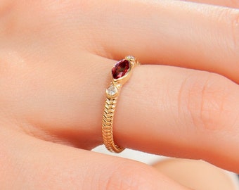 Op de afbeelding: Een gouden ring met een rode edelsteen en kleine witte edelstenen. De ring heeft een gevlochten ontwerp.