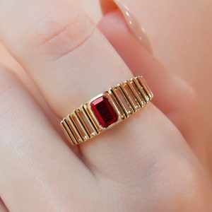 Könnte beinhalten: Ein goldener Ring mit einem roten rechteckigen Edelstein, der in der Mitte eingesetzt ist. Der Ring hat ein einzigartiges Design mit mehreren goldenen Balken, die entlang des Bandes verlaufen.
